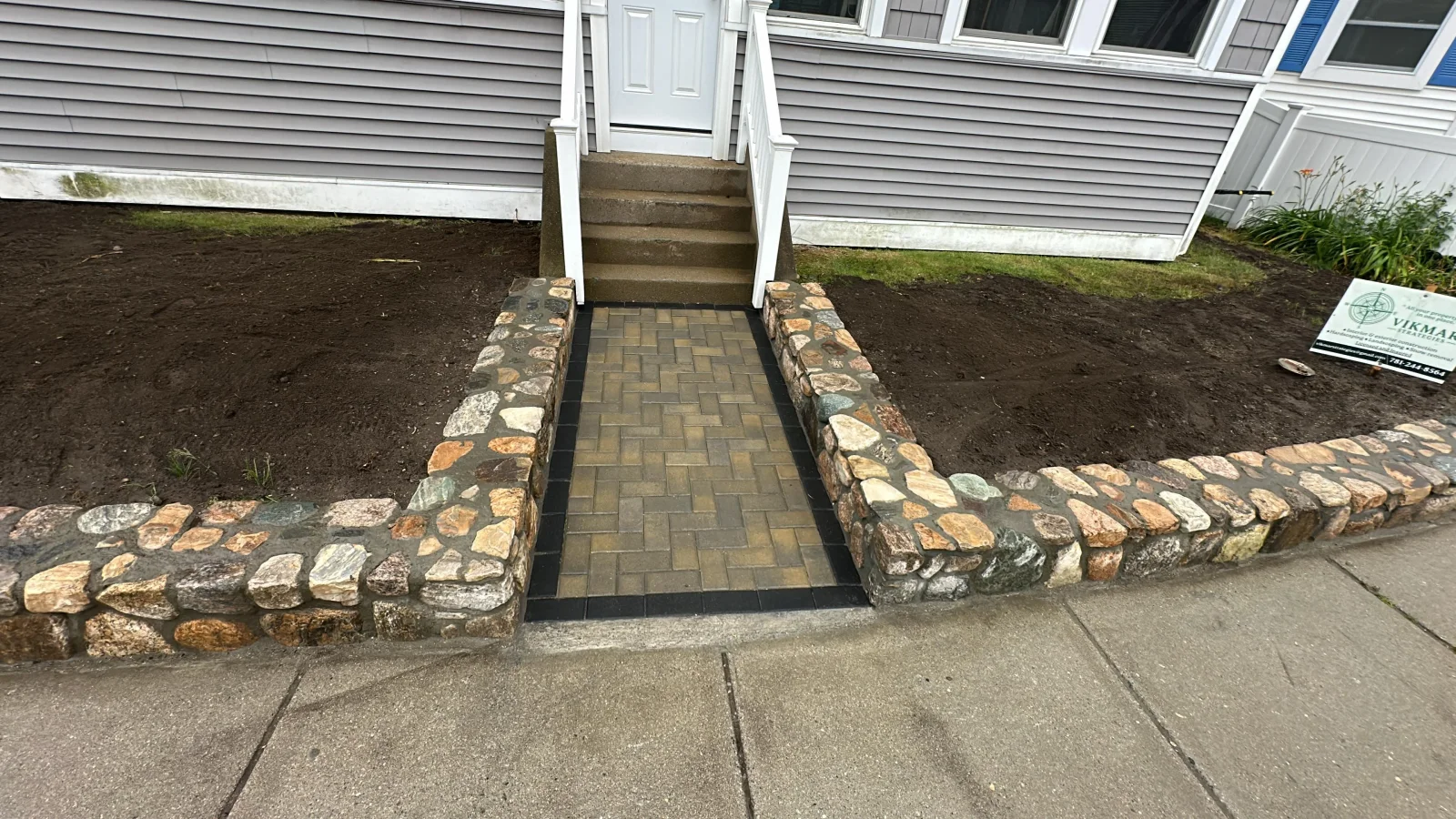 Herringbone brown paver walkway with black border and mixed fieldstone retaining walls leading to front steps