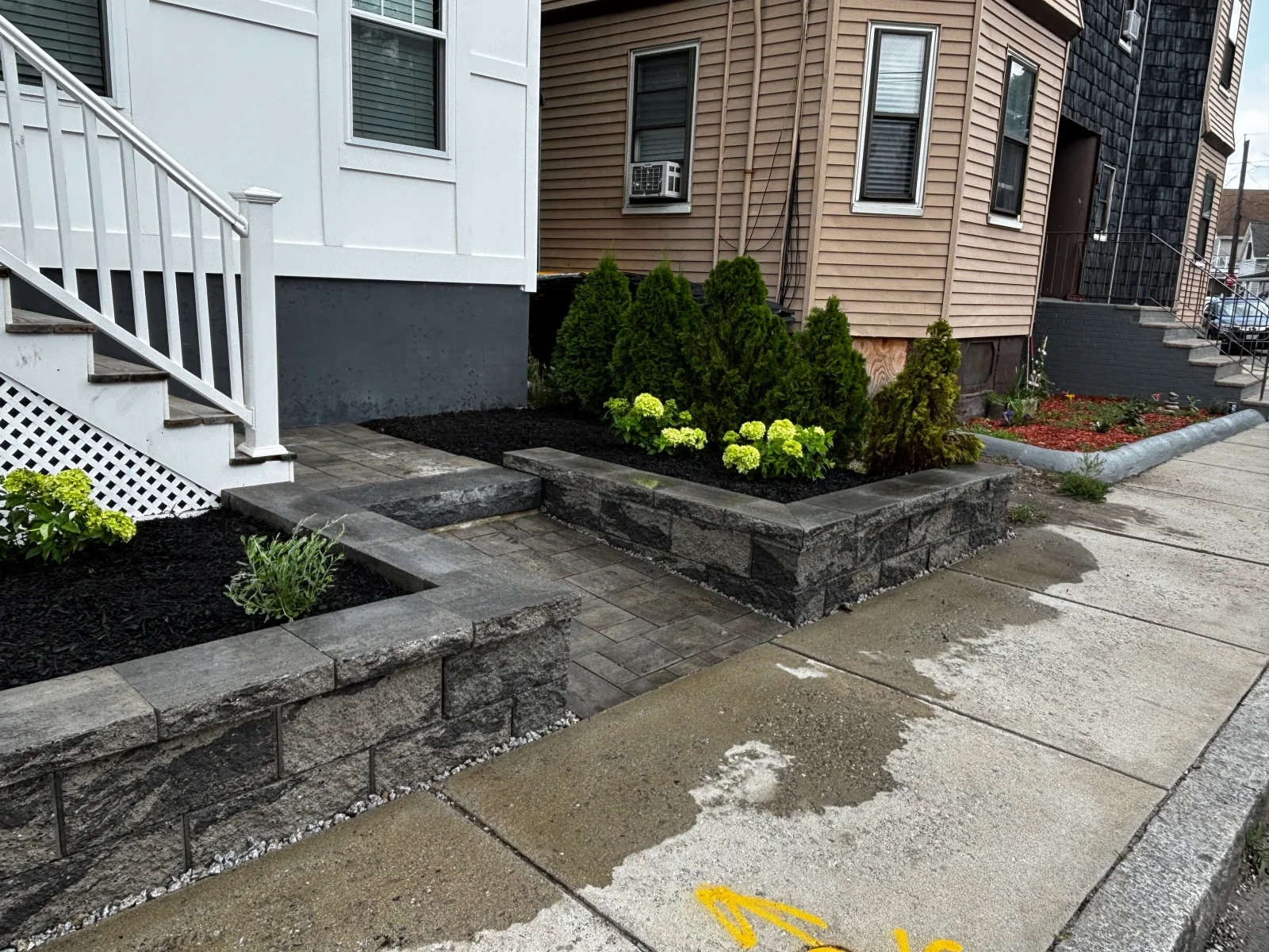 Dark gray segmental retaining walls with paver walkway, black mulch beds, and evergreen and hydrangea plantings beside a front stair