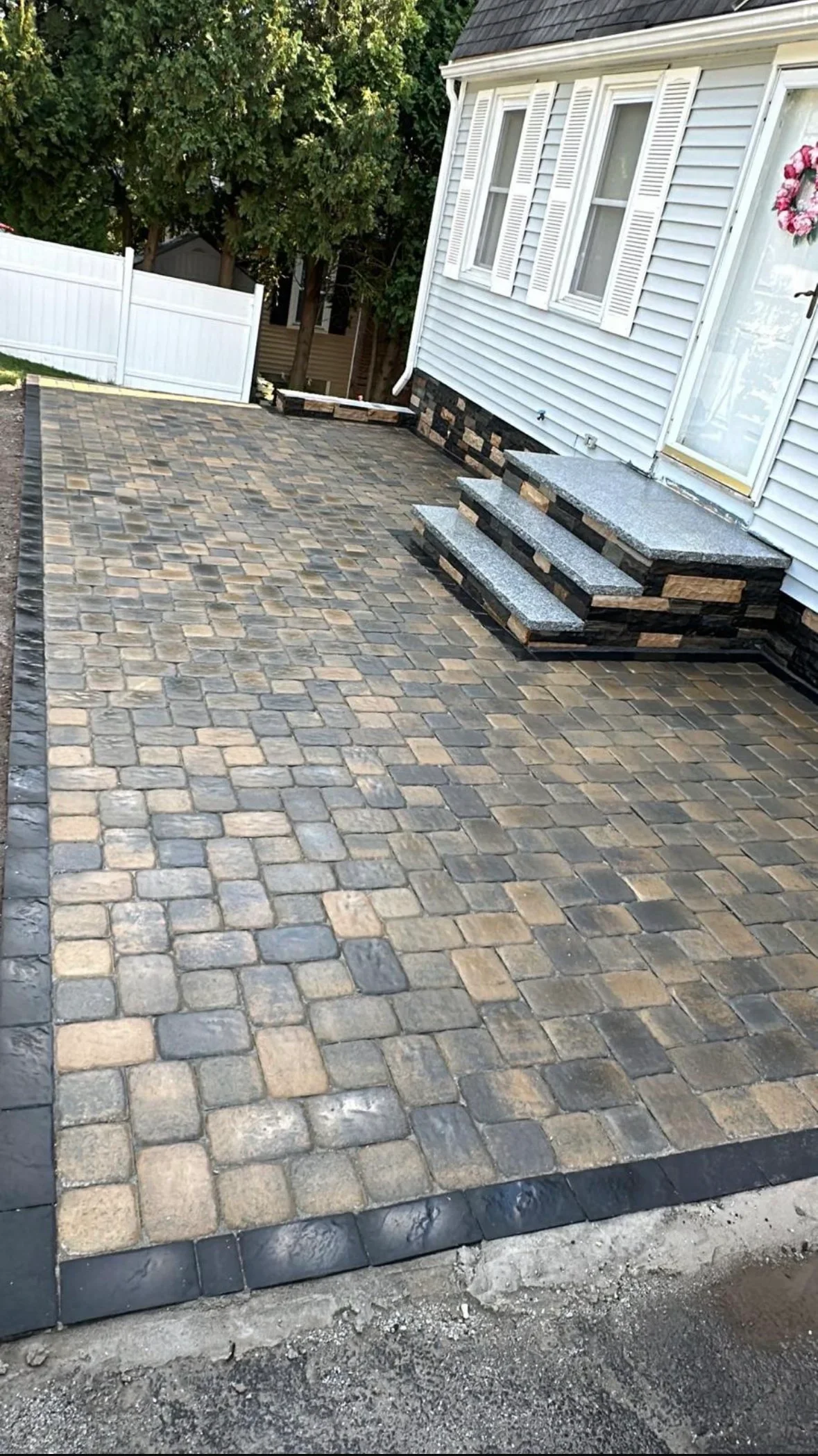 Mixed tan and charcoal paver patio with dark border and gray stone-tread steps beside vinyl-sided home