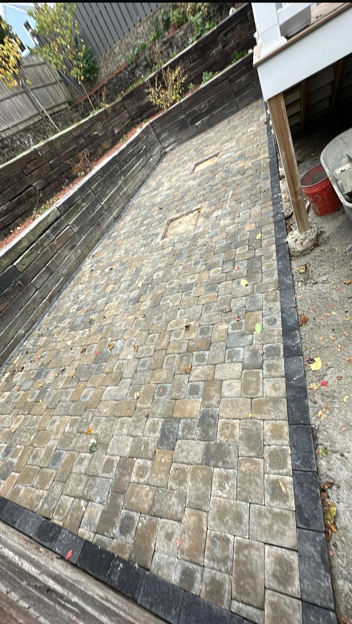 Interlocking tan and gray paver patio with charcoal border beside stacked retaining wall