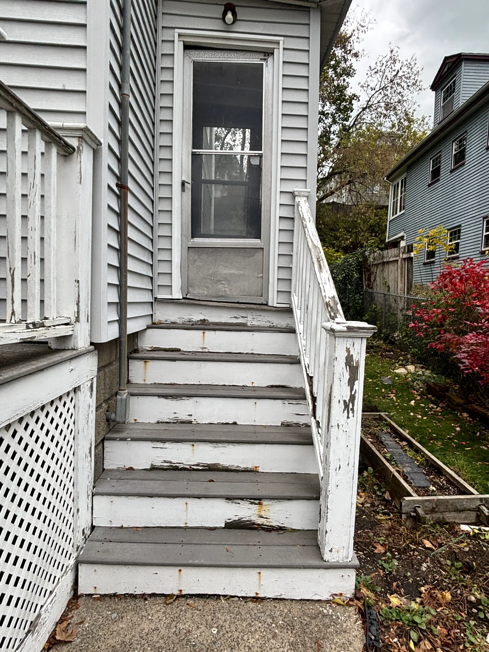 Weathered wood entry steps with peeling white paint and gray treads beside a white wood handrail and screen door