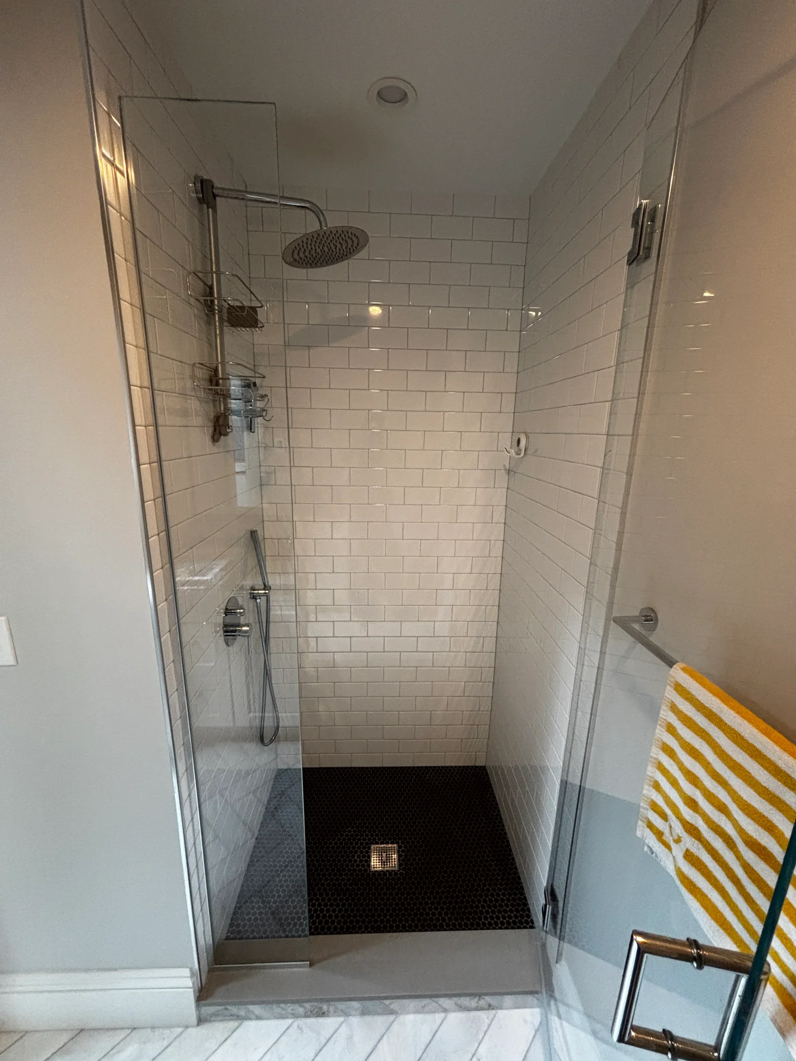 Walk-in shower with white subway tile walls, black hex mosaic floor, chrome rain showerhead, and frameless glass enclosure