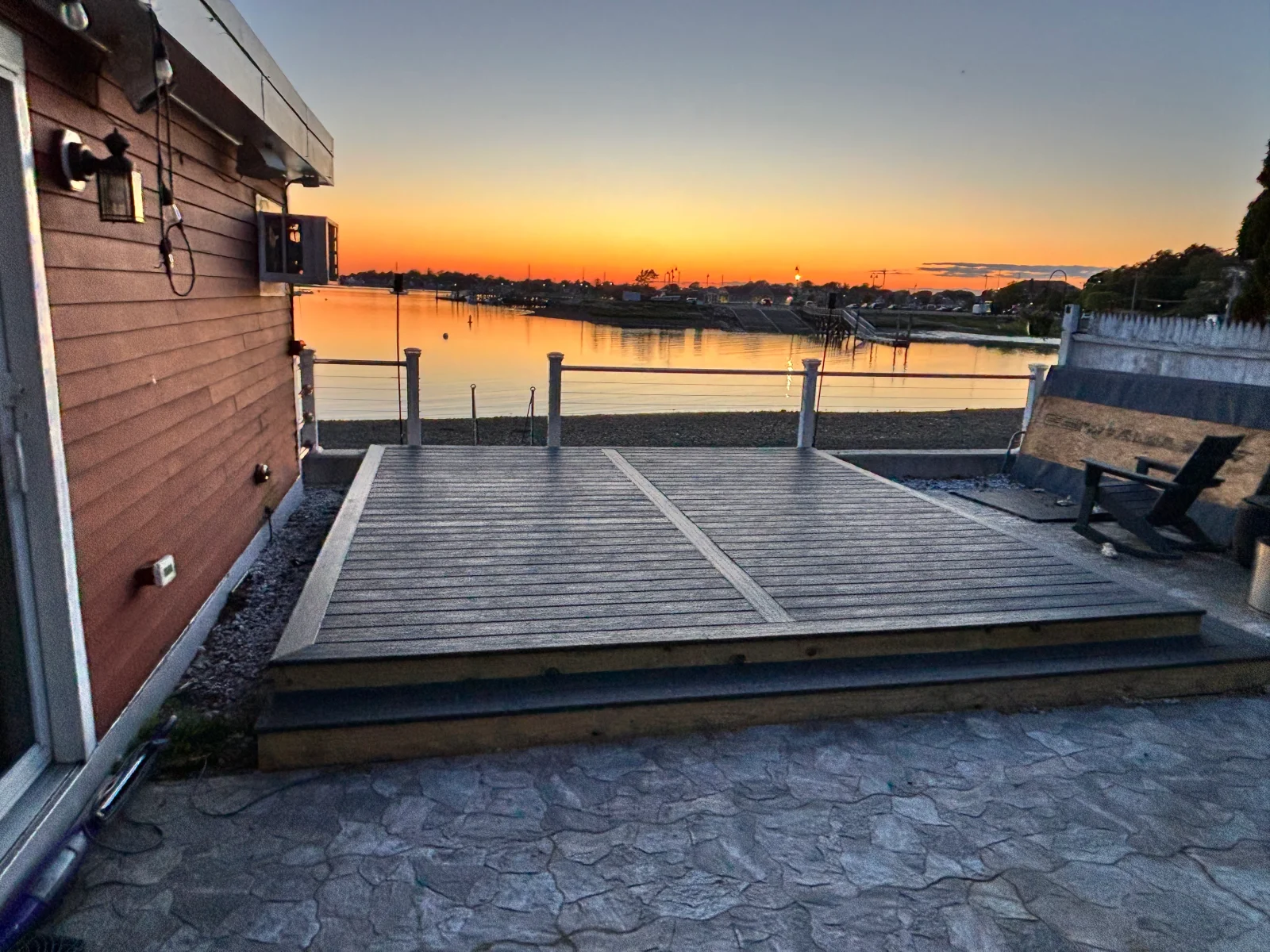 Gray composite deck with cable railing and stamped concrete patio beside red siding at sunset
