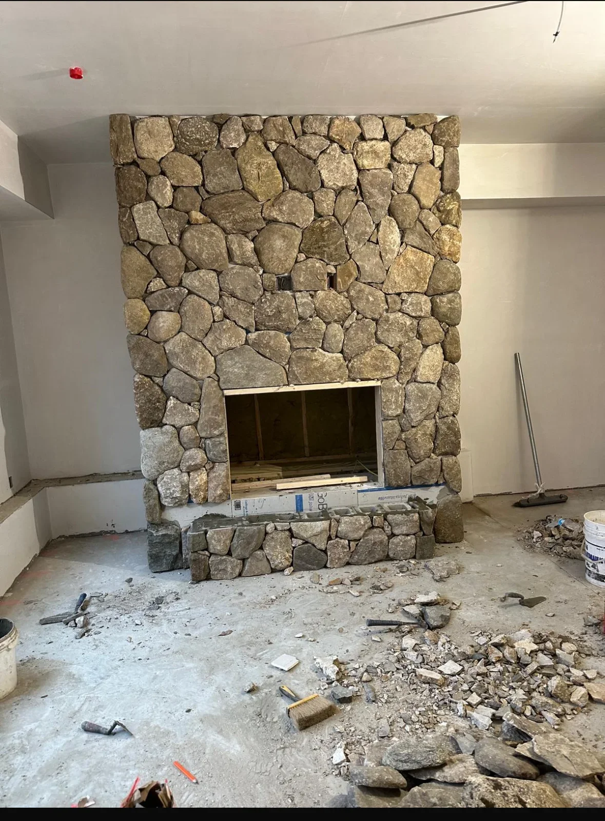 Irregular fieldstone fireplace surround under construction with firebox opening, stone hearth, and masonry debris on concrete floor