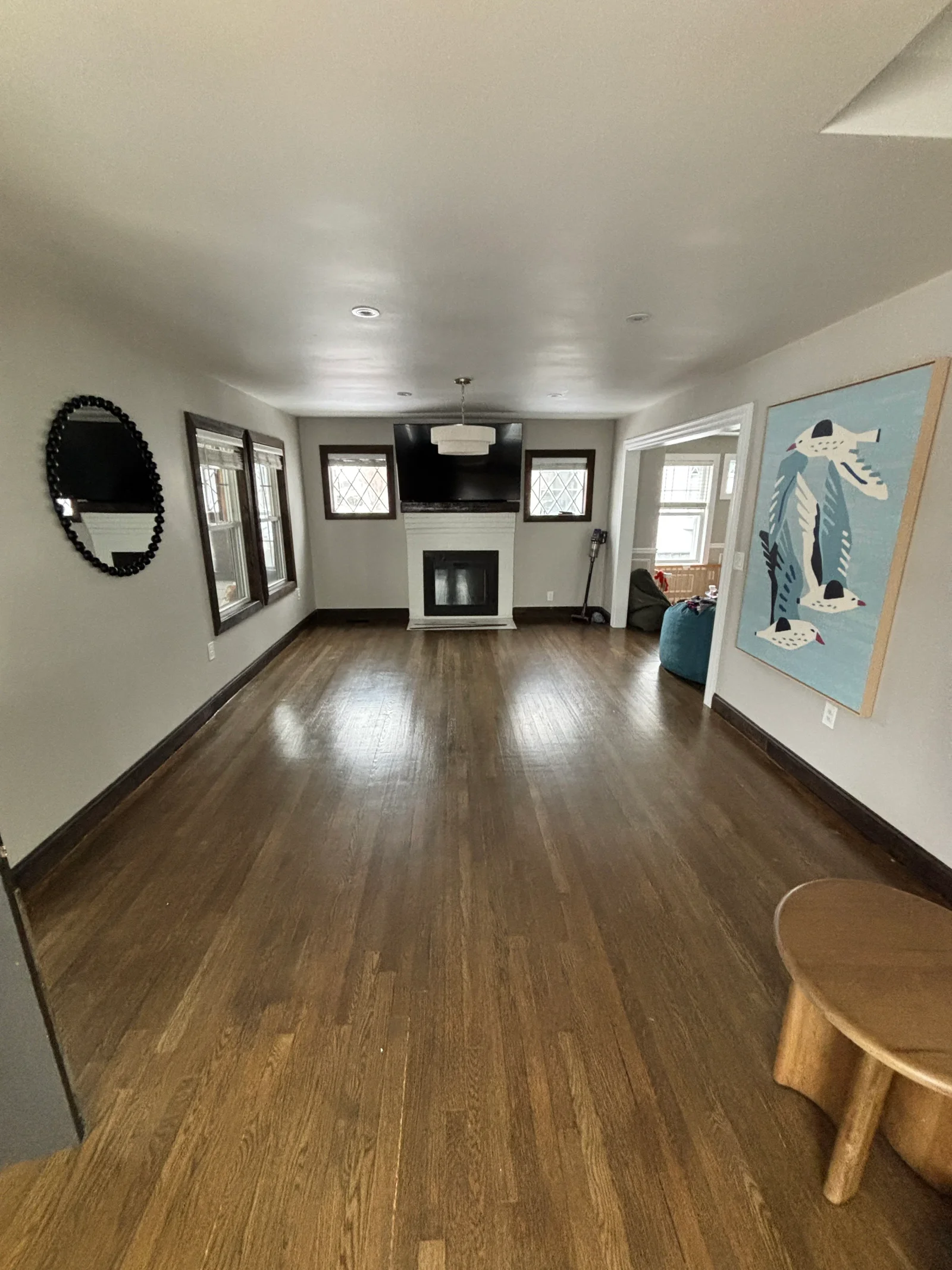 Living room with refinished medium-brown hardwood floors, dark stained trim, white painted fireplace surround, and light gray walls