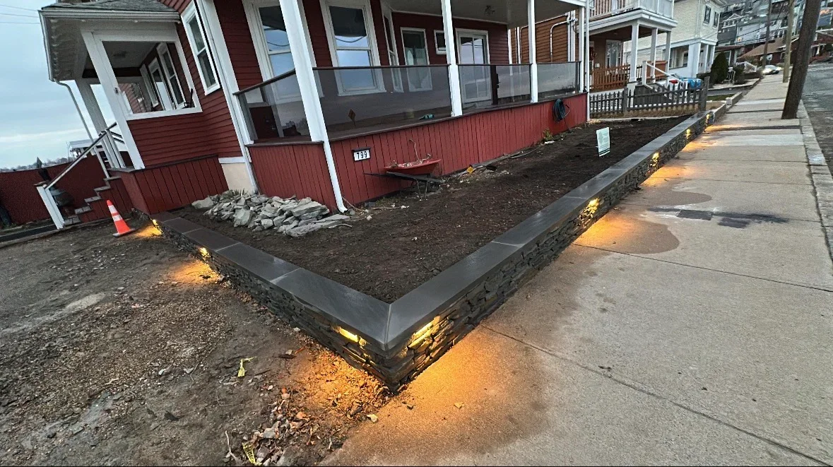 Stacked stone retaining wall with dark gray cap stones and recessed warm landscape lighting beside a sidewalk