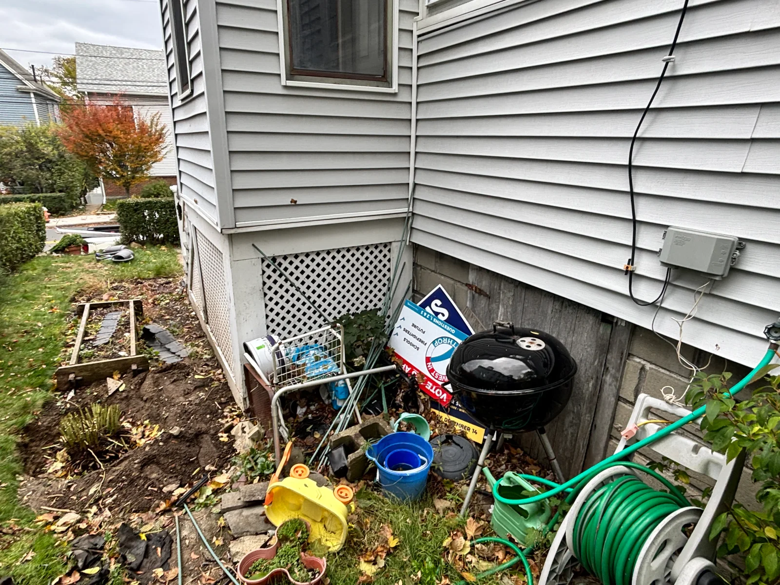 Gray vinyl siding, white lattice foundation vent, and cleared soil beside a timber-framed garden bed